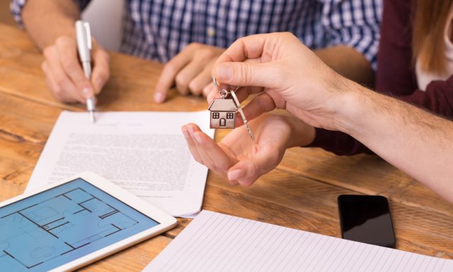 Happy young couple getting keys of their new house. Close up of female hand receiving keys from house broker. Closeup of the hand of a real estate agent who give the house keys to a woman while her boyfriend signing a contract.